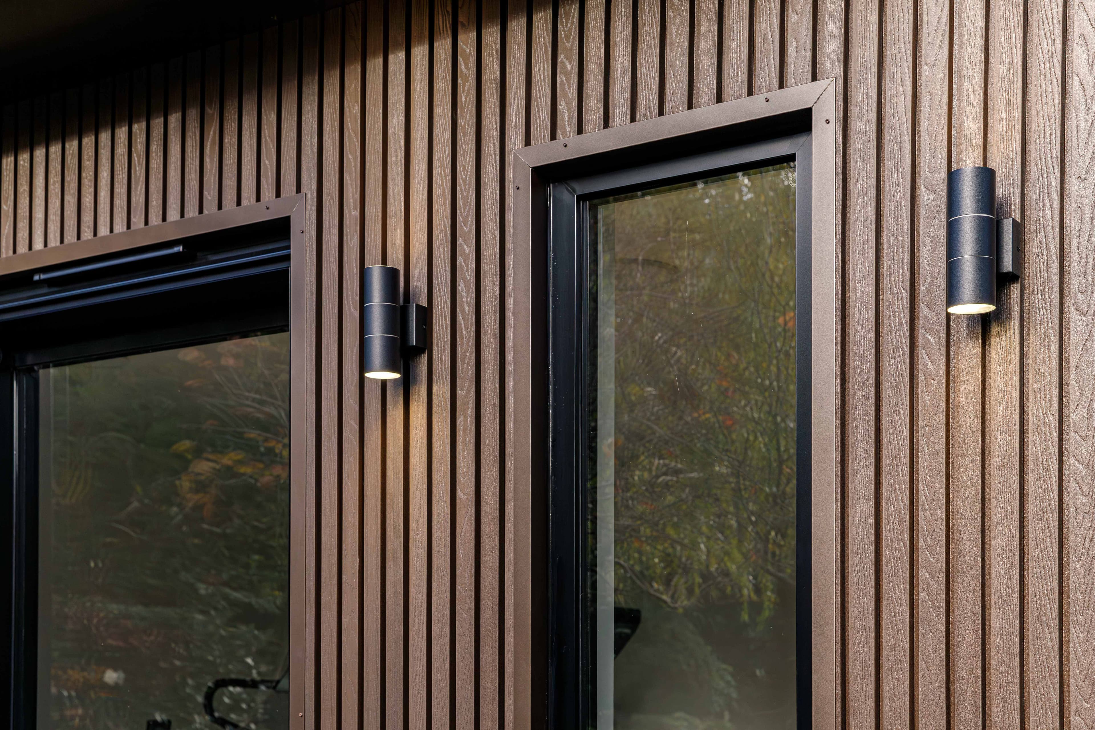 Close-up of brown composite wood-grain cladding on a Bedfordshire garden room with black up-and-down wall lights beside the windows
