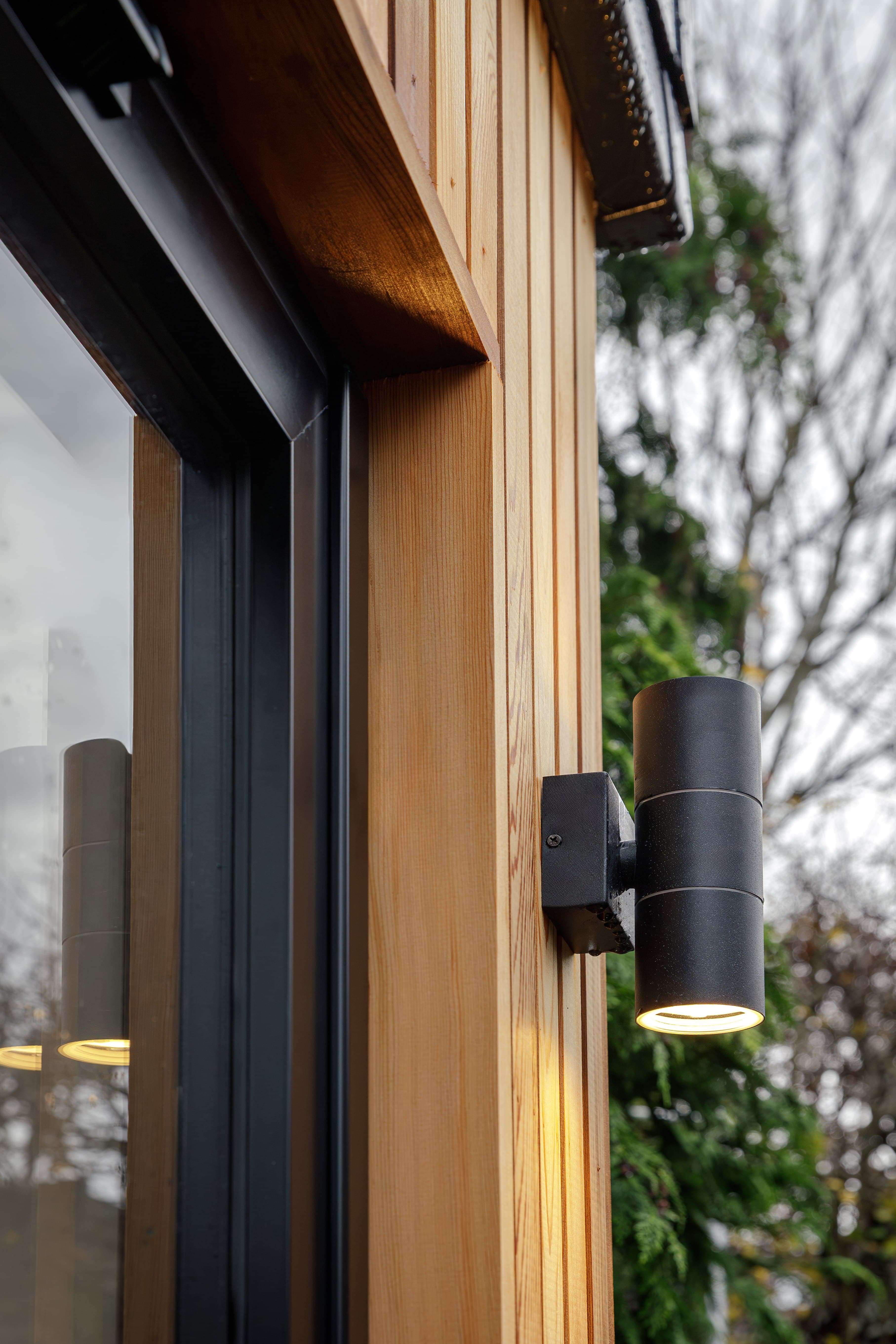 Close-up of a black up-and-down wall light on natural cedar cladding beside a black-framed window of a Bedfordshire garden room
