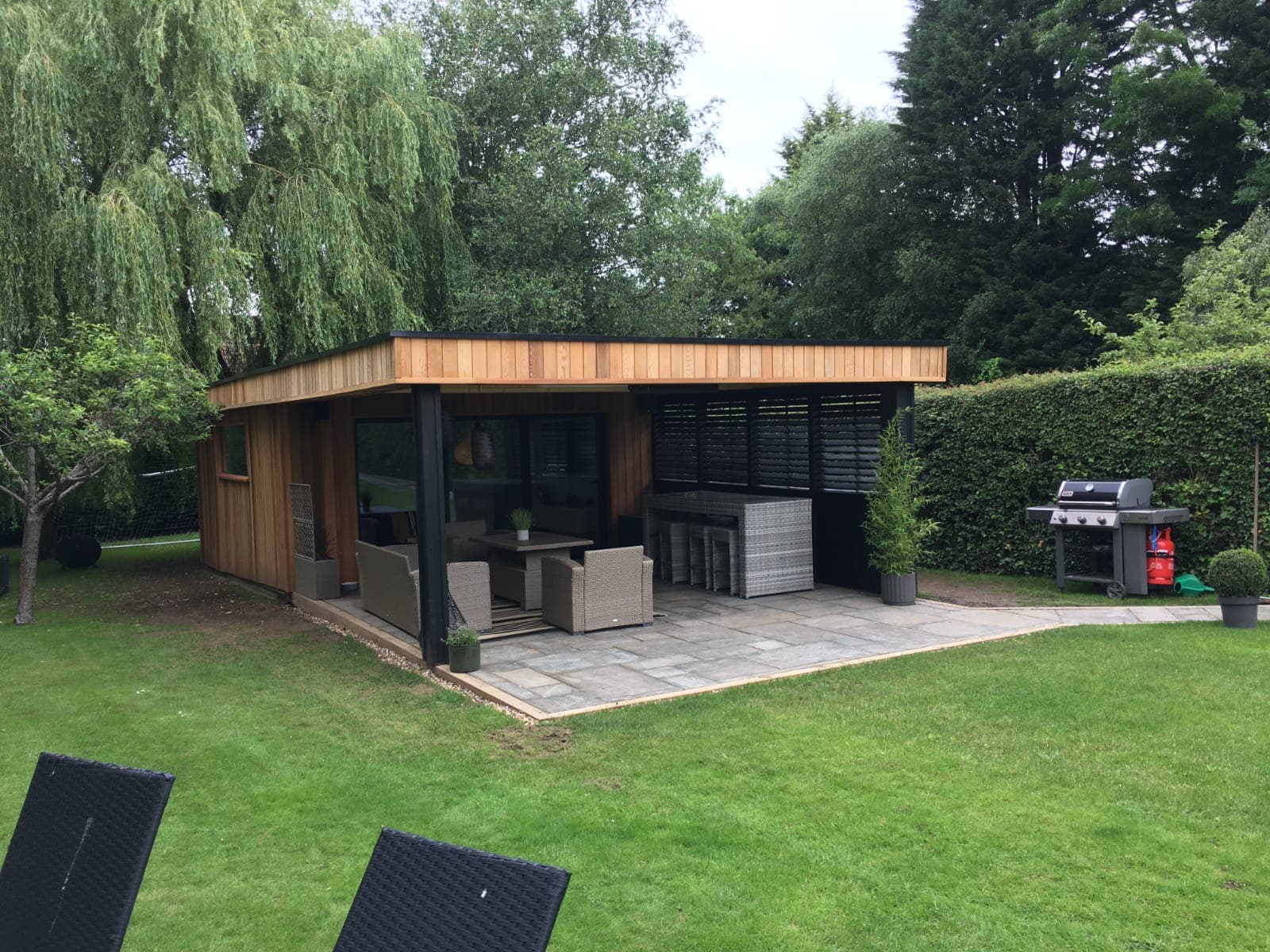 Cedar and black-clad garden room in Bedfordshire with a covered terrace, rattan dining set and barbecue on the lawn