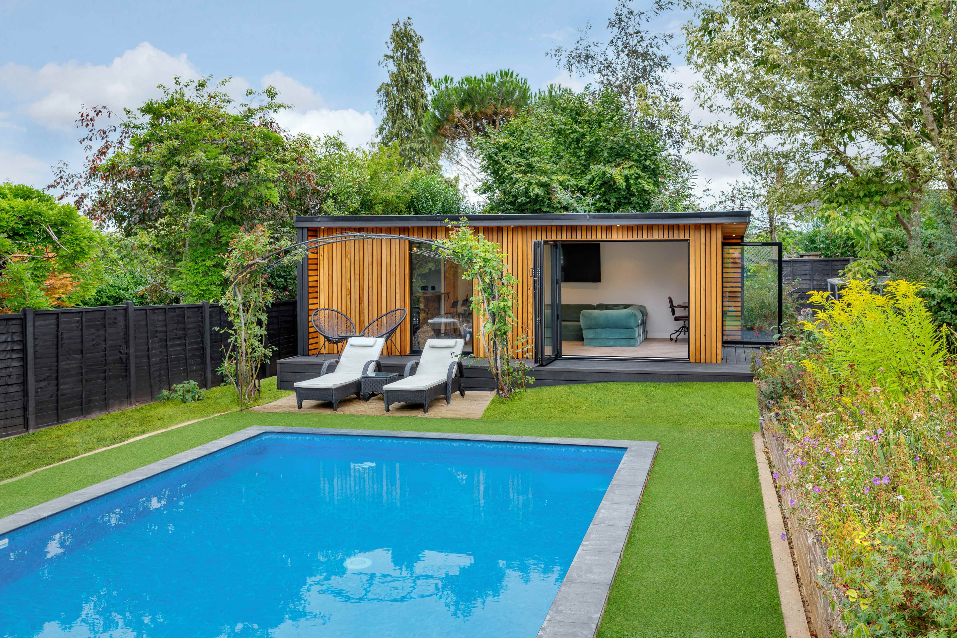 Cedar-clad garden room in Bedfordshire beside a swimming pool with sun loungers and a sofa inside