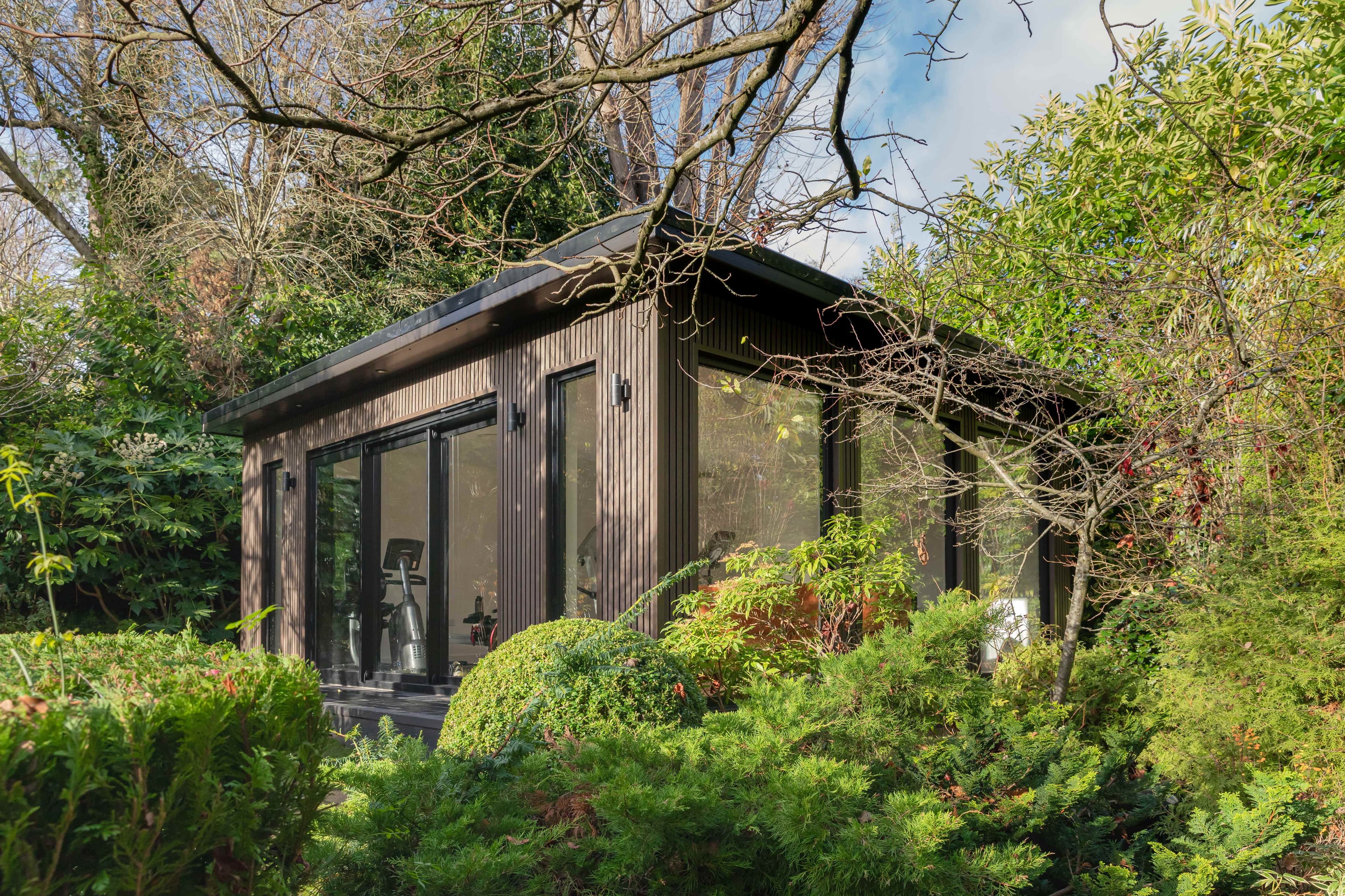 Dark-clad garden gym in Bedfordshire set among mature trees and shrubs with exercise equipment inside