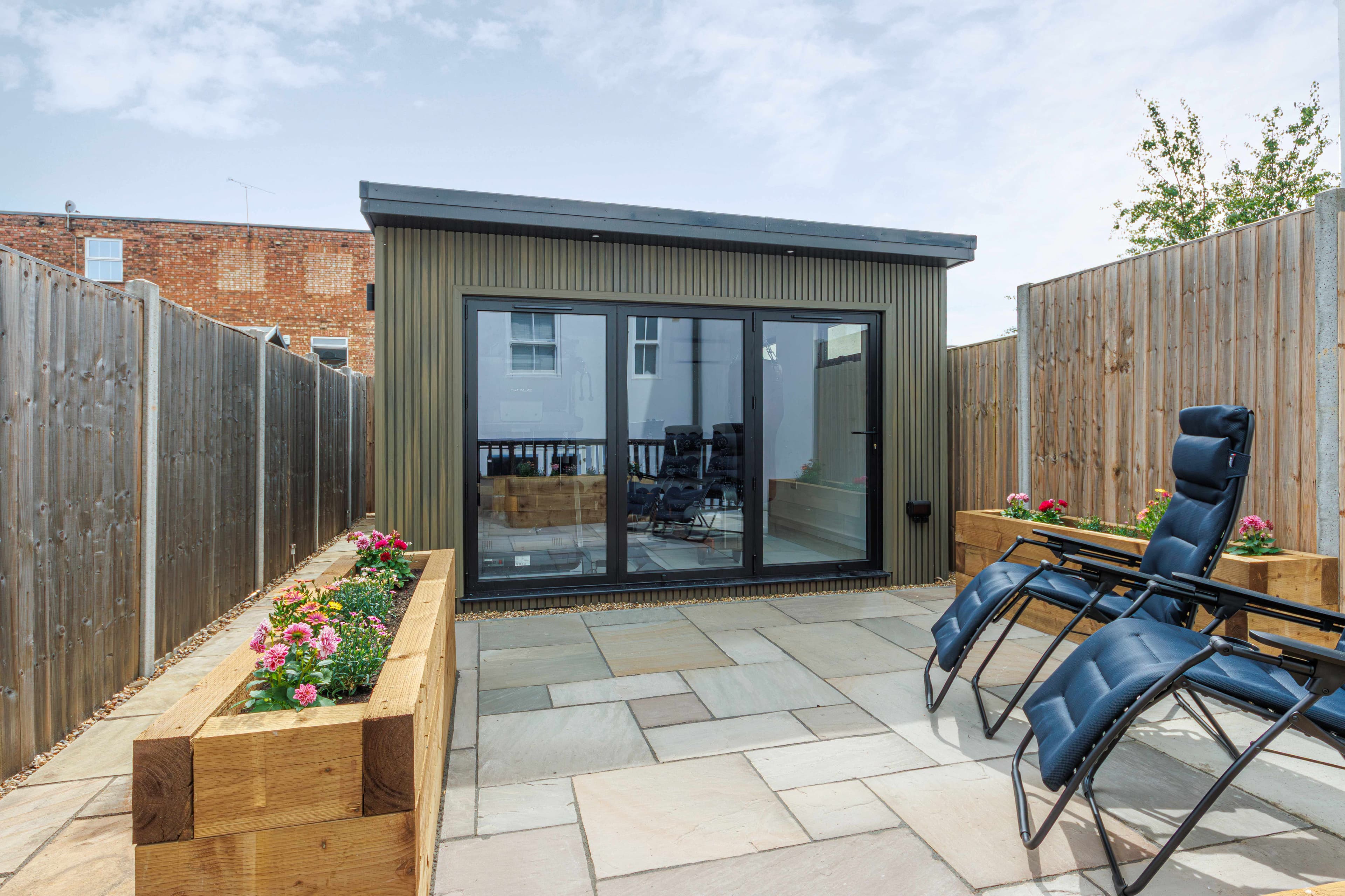 Green-clad garden room in Bedfordshire with anthracite bifold doors, a sandstone patio, raised flower beds and reclining loungers