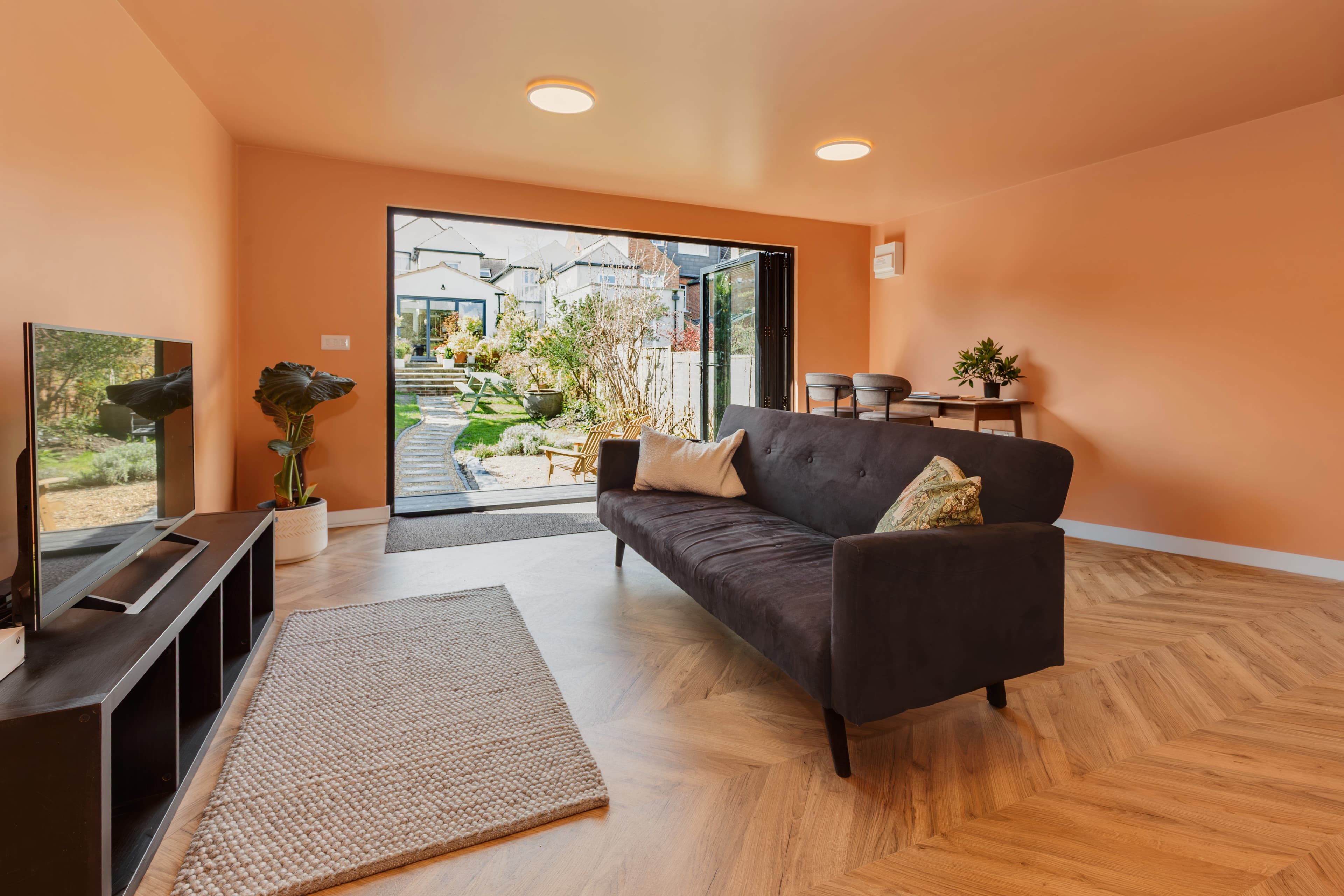 Garden room lounge in Bedfordshire with warm orange walls, a dark sofa and bifold doors open to the garden