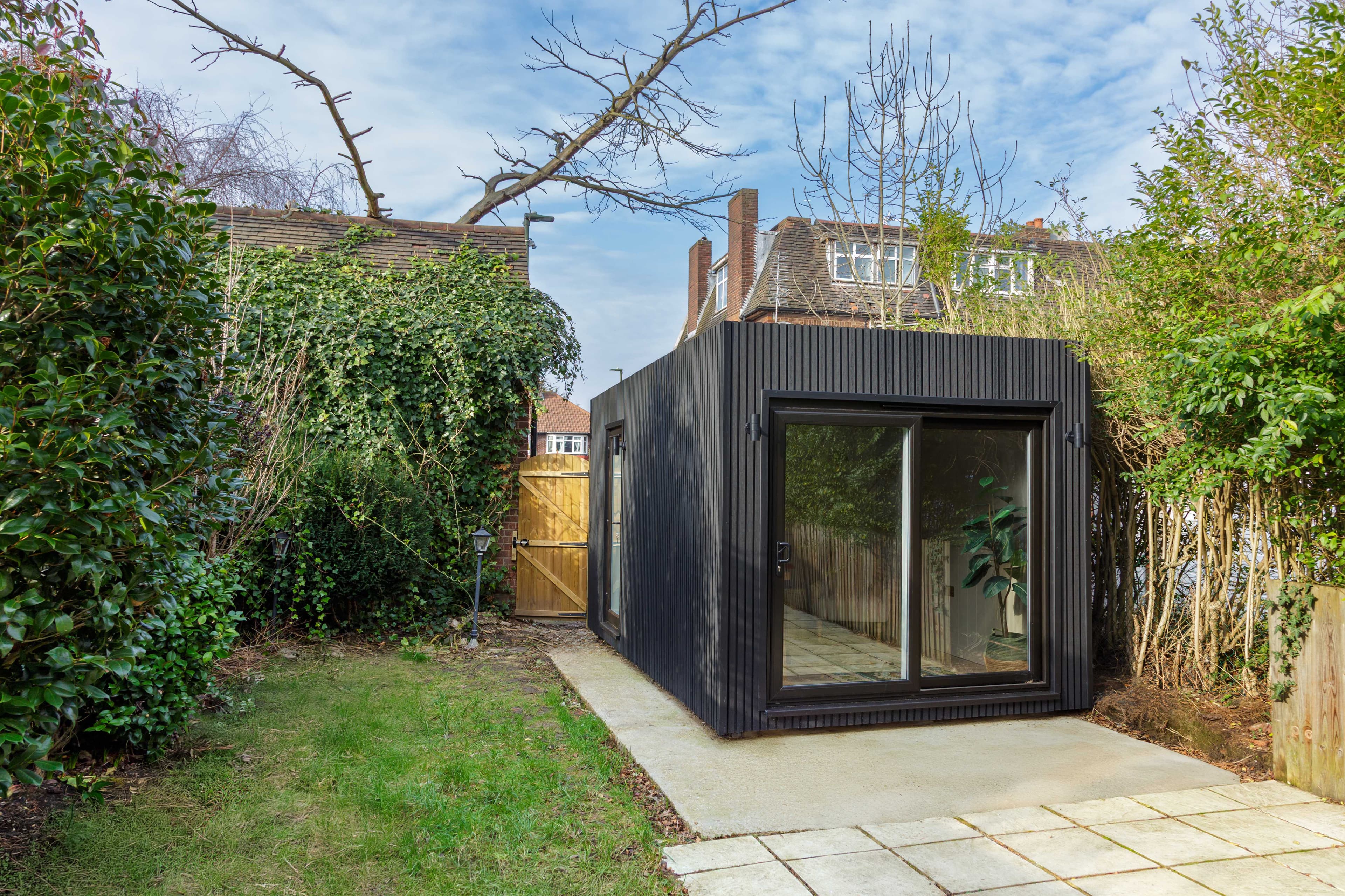 Black slatted garden room in Bedfordshire with a sliding door, set on a sandstone patio between mature hedges