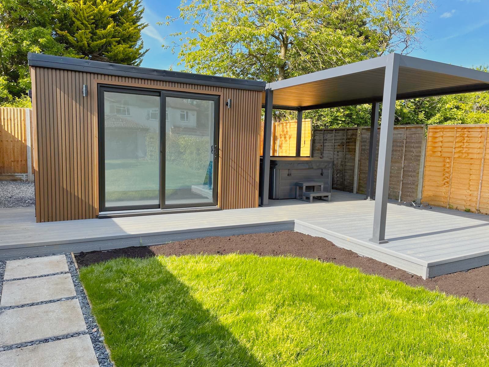 Brown-clad garden room in Bedfordshire joined by a pergola over a hot tub on a grey composite deck