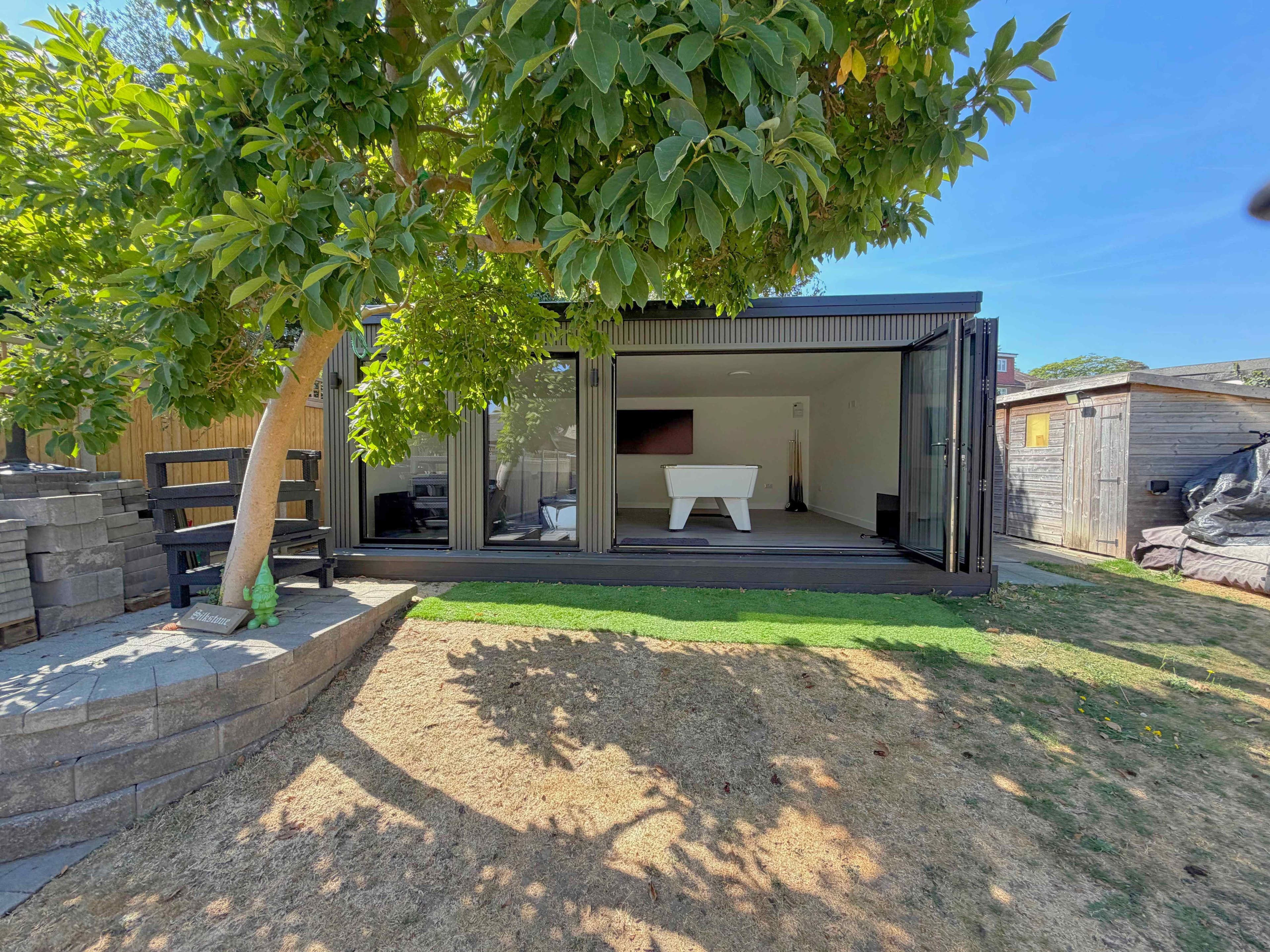 Charcoal corrugated-clad garden games room in a Bedfordshire garden with bifold doors folded back, revealing a white pool table and wall-mounted TV under a leafy tree