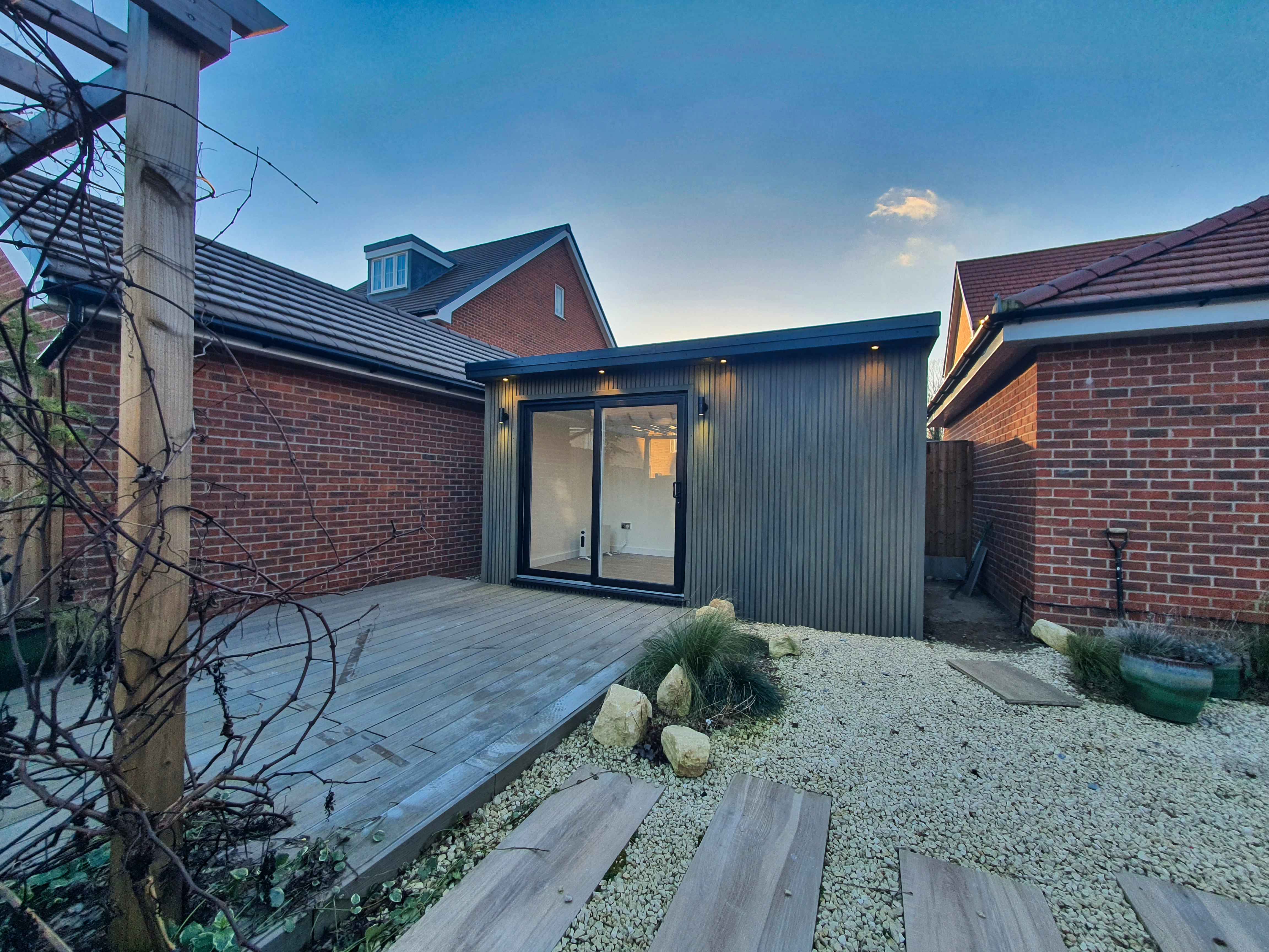 Charcoal-clad garden room in Bedfordshire lit warmly from within at dusk, with a timber deck and gravel path