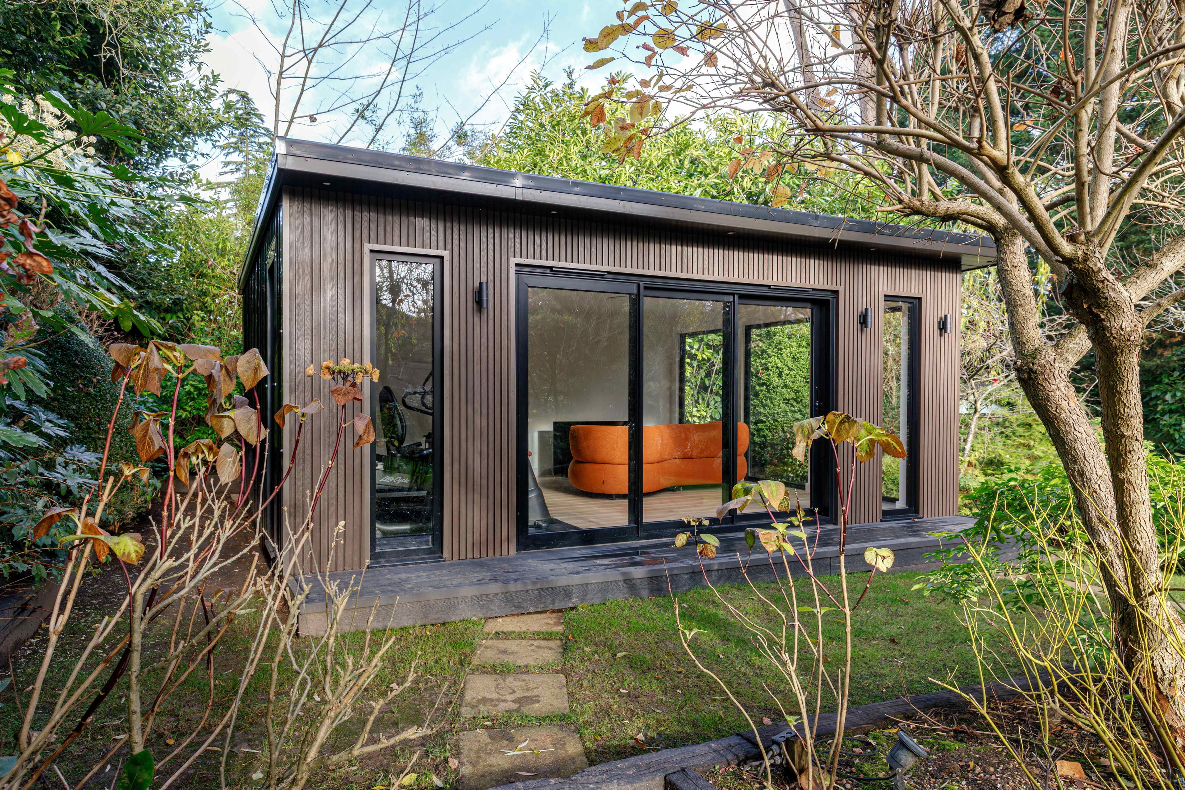 Dark timber-clad garden room in Bedfordshire with wraparound glazing and an orange curved sofa inside, set among woodland planting