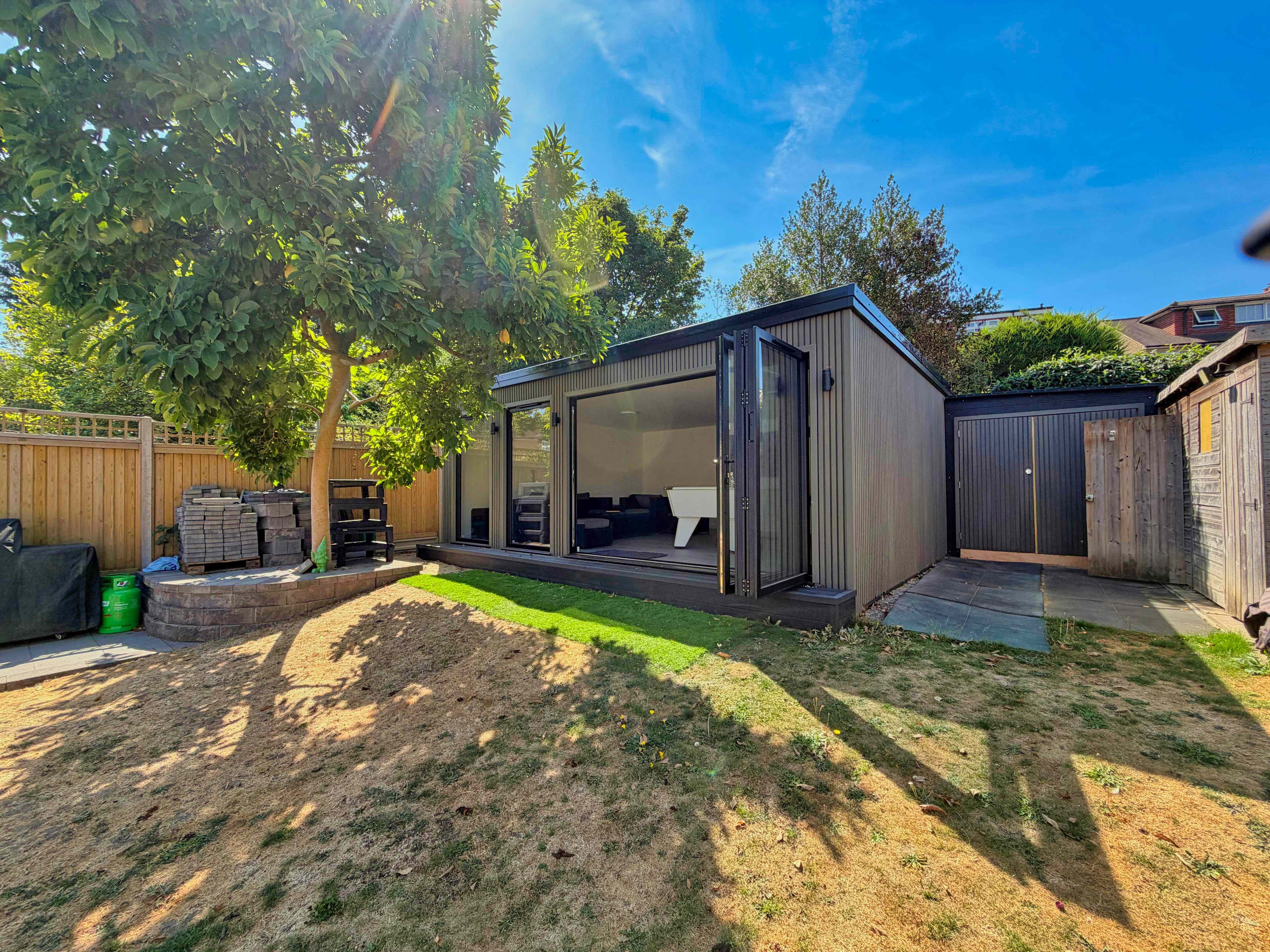 Graphite corrugated-clad garden games room in a Bedfordshire garden with bifold doors open to a white pool table, beside a matching storage unit under a leafy tree