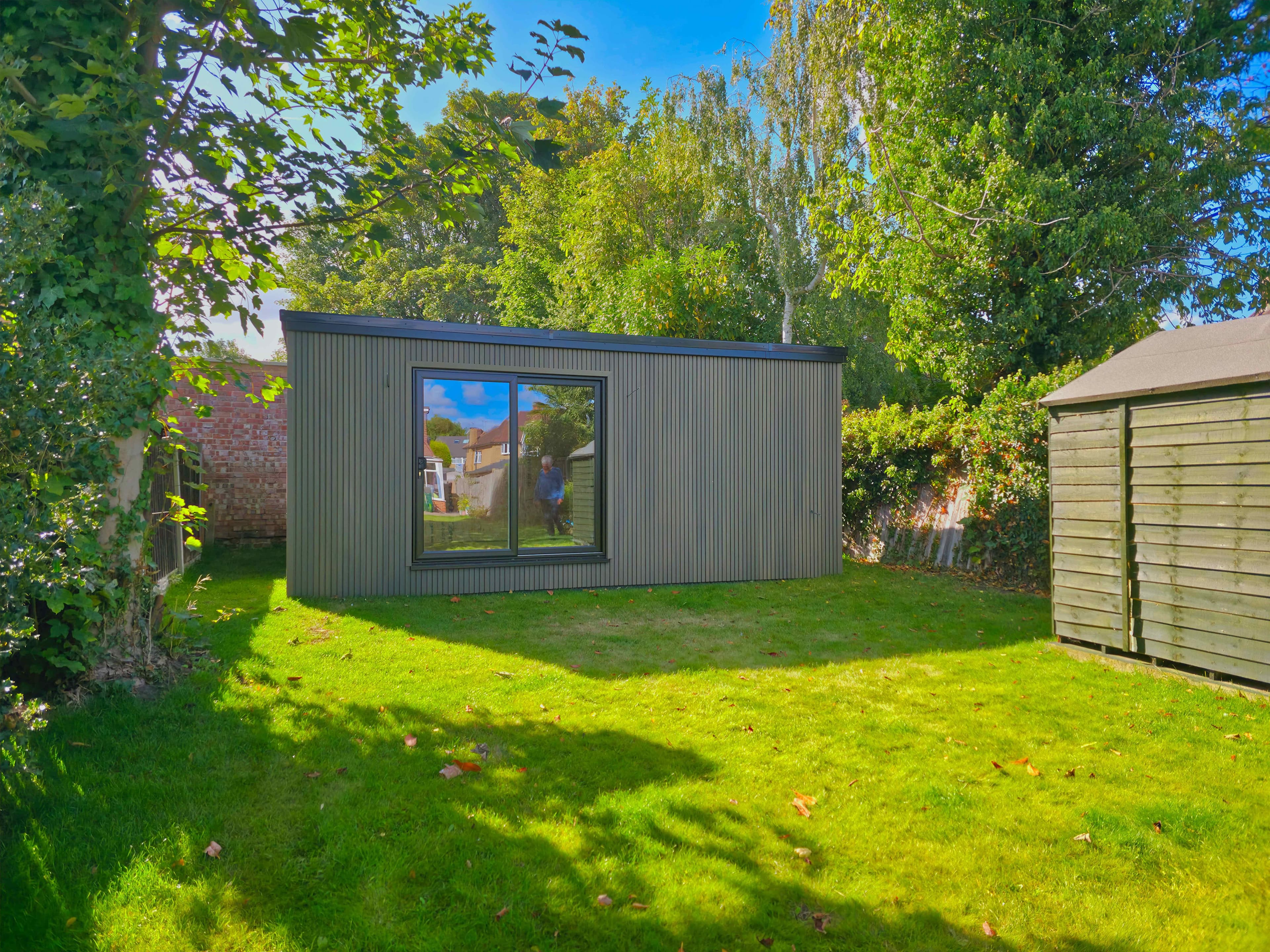 Grey slatted garden room in Bedfordshire with a large sliding window, set on a tree-lined lawn beside a timber shed