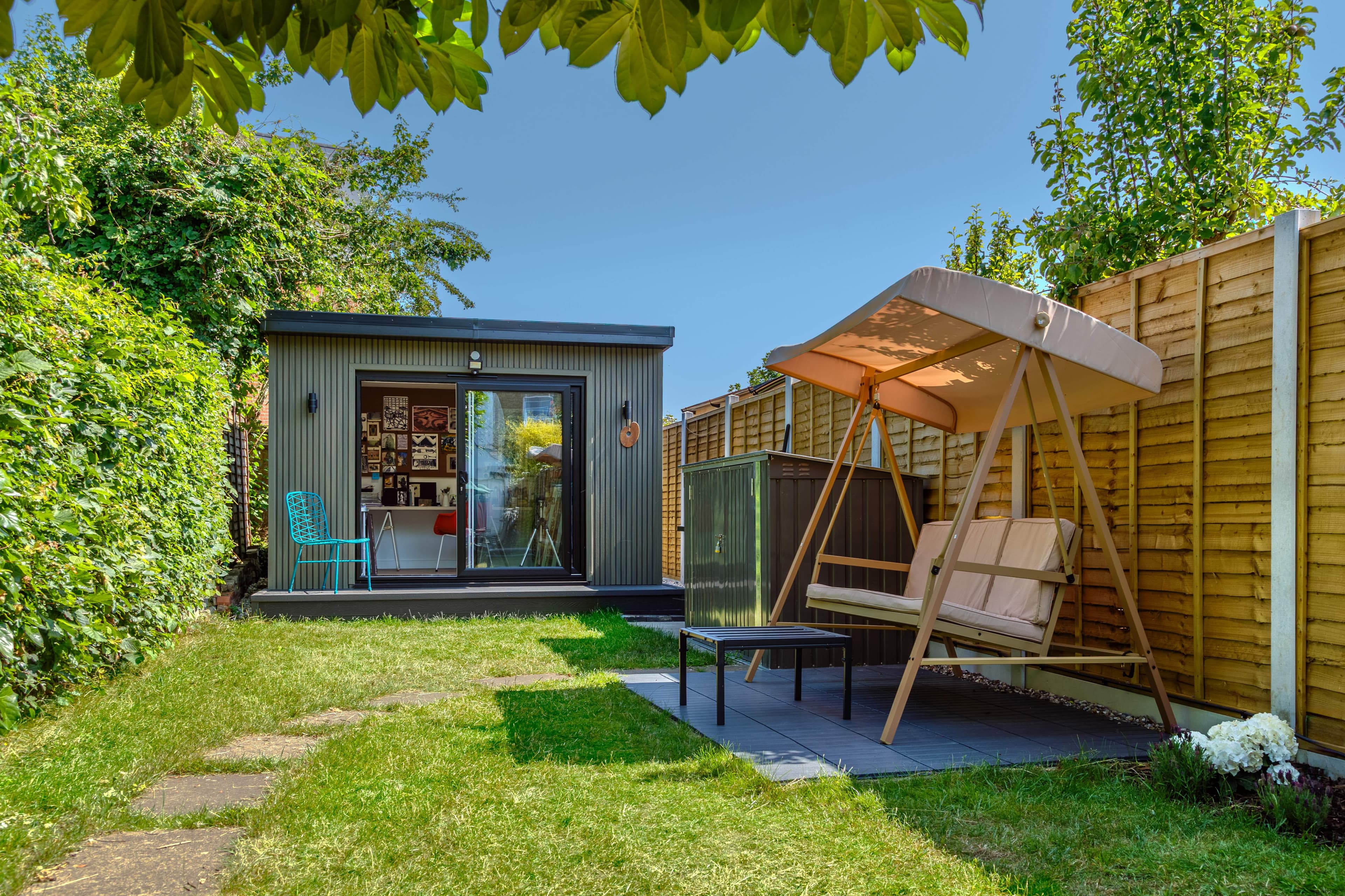 Olive-grey garden art studio in Bedfordshire with a sliding door open beside a canopied garden swing seat